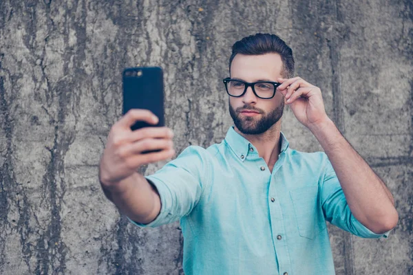 スマート フォンのフロント カメラで Selfie を撮影する夢のような式で顔にメガネのアイレットを保持している過酷な残忍な男の肖像は 灰色の石の壁の外に分離 アプリ電子デバイスの概念 — ストック写真