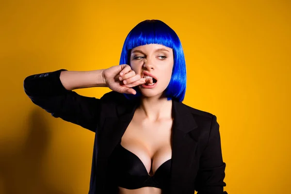 Portrait of harsh brutal woman in black jacket bright blue wig with big tits biting forefinger looking to the side isolated on yellow background. Holiday theme party dress-code concept