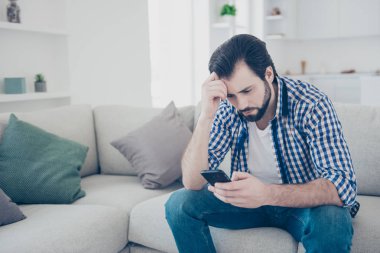 Portrait of tired alone serious man sitting indoor using smart phone, internet, checking email, having problem in relationship chatting with his girlfriend worry about bad news clipart