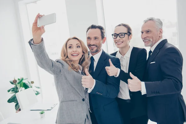 魅力的なスタイリッシュなうれしそうな 自信を持って事業者が職場 ワークステーション フロント カメラで撮影 Selfie に立って 楽しいを持っているスマート フォンを使用して表示を親指指の手で — ストック写真