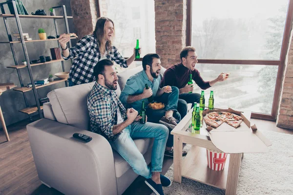 Four attractive, crazy, stylish, bearded guys with modern hairstyle watching match on television, together sitting in livingroom, having lager and snacks, yelling, cheering for their favorite team