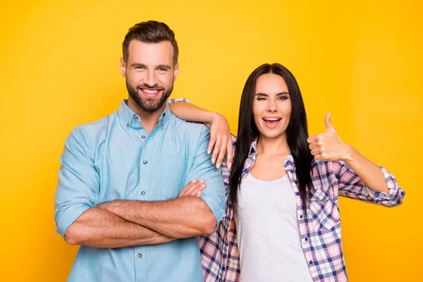 Portrait of comic couple in casual clothes, handsome man holding arms crossed, beautiful woman gesturing thumb up and winking with one eye isolated on bright yellow background — Stock Image Portrait Comic Couple Casual Clothes Handsome Man Holding Arms Crossed Stock Photo