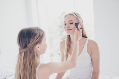 Preteen schoolgirl style fun blonde long straight hair concept. Close up portrait of cute glad kid doing makeup for her older adopter sister applying maquillage on cheekbones holding brush in hand clipart