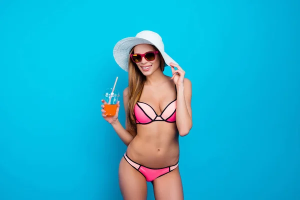 Portrait of trendy stylish girl in summer glasses swim wear holding hat and alcohol beverage with tubule standing over blue background