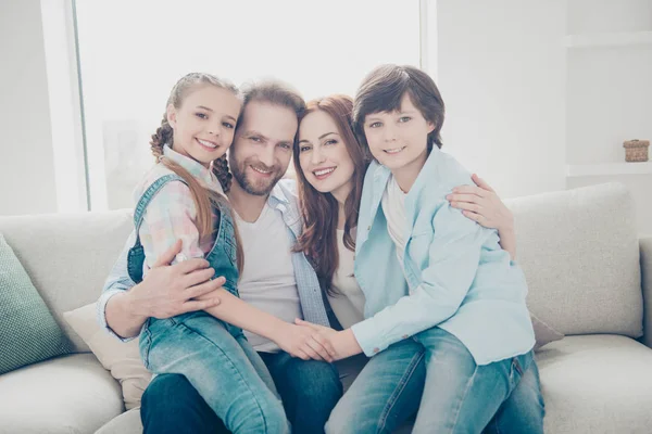 Portrait of cheerful full family with two kids sitting on parent\'s legs indoor in living room bonding holding hands looking at camera. Upbringing care concept