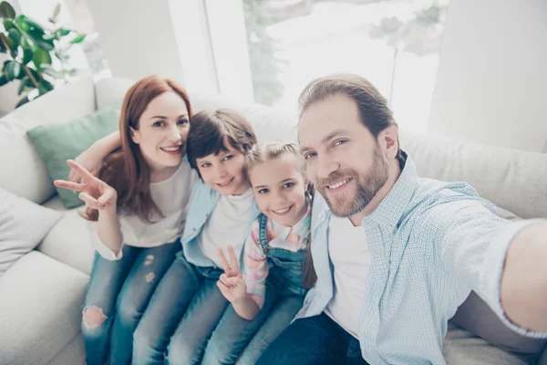 High angle view self portrait of friendly lovely family with two kids embracing sitting on sofa shooting selfie on front camera having video-call gesturing v-sign hi symbol