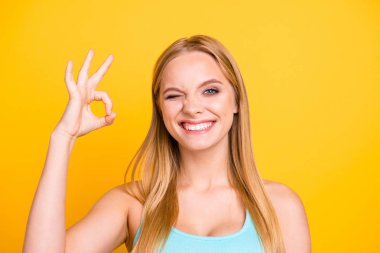 I like it! Close up portrait of young, gorgeous and charming girl isolated on yellow background, in trendy wear, with long blond hair. Female model wink and showing ok symbol clipart