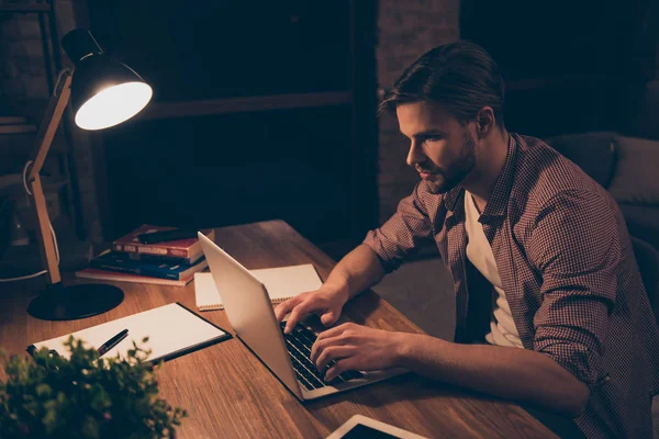 Portrait of attractive programmer, hard worker, busy man in shirt with hairstyle working at night, taking work at home, looking at screen of laptop, sitting in work place, holding hands on keypad
