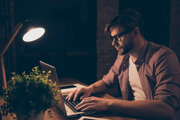 Portrait of attractive programmer, hard worker, busy man in shirt with hairstyle working at night, taking work at home, looking at screen of laptop, sitting in work station, holding hands on keypad