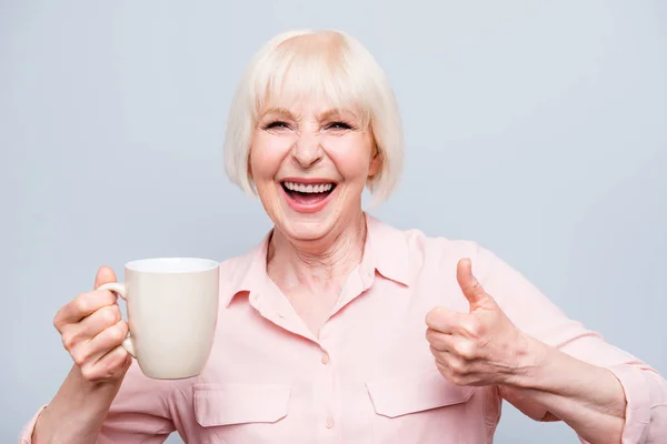 Portrait of old excited lady showing thumbs up gesture, smiling laughing, holding cup drinking coffee, tea, beverage on grey background, isolated