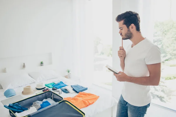 It is better to write everything! Young bearded man standing in a bright bedroom in front of clothes thinks what else needs to be folded into a suitcase before traveling