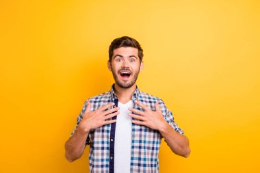 I won?! Young brunette man with an enthusiastic face look into the camera put his hands on his chest and wide open mouth and eyes with surprise isolated on shine yellow background clipart