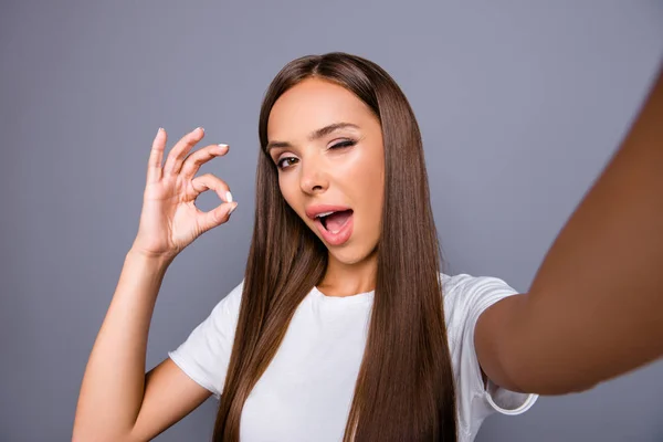 Imagen de cerca de atractiva morena mujer guiño con la boca abierta disparando selfie, gesto ok signo con los dedos, tener videollamada, de pie aislado sobre fondo gris — Foto de Stock