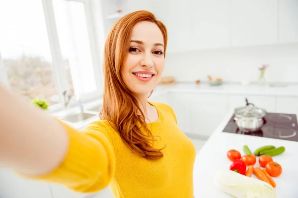 Mujer joven en suéter amarillo hacer selfie en la cámara frontal del teléfono inteligente contra el tomate, pepino, pimienta, col, zanahorias para el desayuno en la mesa en el interior de la luz moderna —  Fotos de Stock