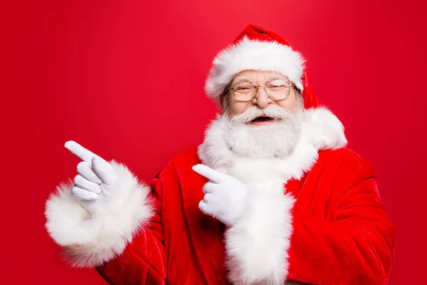 时髦的喜悦时尚的圣诞老人在服装手套头饰和眼镜与白胡子指示撇开食指两手展示展示产品推广隔离在诺红背景上 — 图库照片