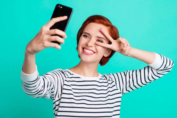 Close up portrait of cheerful ginger attractive readhead she her lady with telephone in hand take self photos making v-sign near eye say hi wearing white striped sweater isolated on teal background