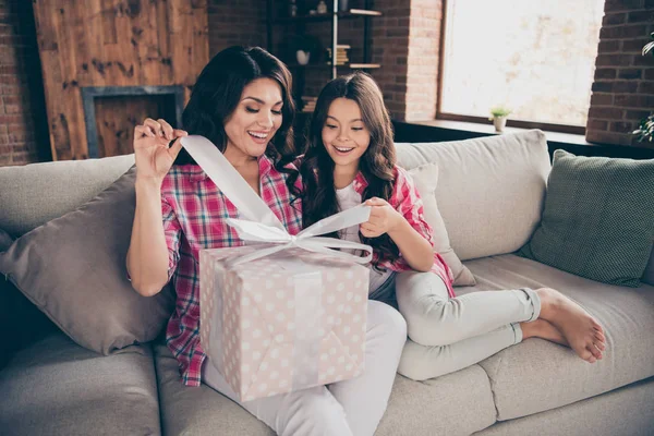 Portrait of two nice cute winsome attractive lovely lovable charming cheerful cheery people wearing checked shirt opening large bow ribbon box sitting on divan indoors — Stock Photo, Image