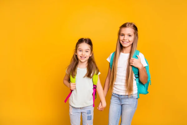 Portrait of two nice-looking attractive lovely cheerful positive pre-teen girls with backpacks holding hands isolated over bright vivid shine yellow background — Stock Photo, Image