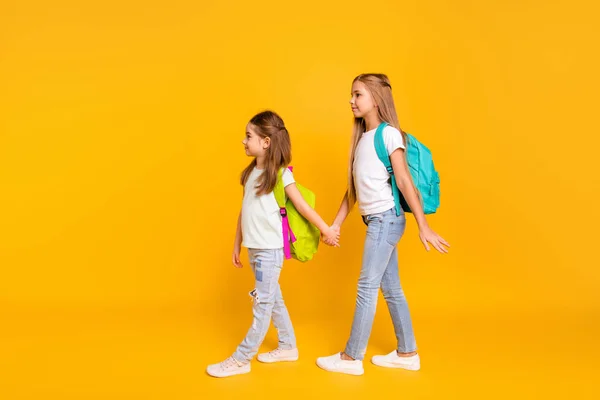 Full length body size profile side view portrait of two nice attractive confident smart pre-teen girls with colorful backpacks holding hands isolated over bright vivid shine yellow background — стоковое фото
