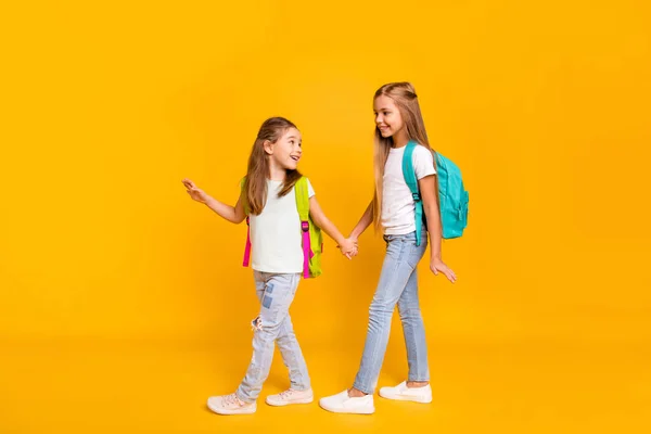 Πλήρες μήκος σώματος μέγεθος προβολής δύο ωραία ελκυστικά χαρούμενα έξυπνο έξυπνο προ-έφηβος κορίτσια με πολύχρωμα σακίδια, κρατώντας τα χέρια πίσω στο σχολείο απομονωμένη φωτεινή λάμψη ζωηρό κίτρινο φόντο — Φωτογραφία Αρχείου