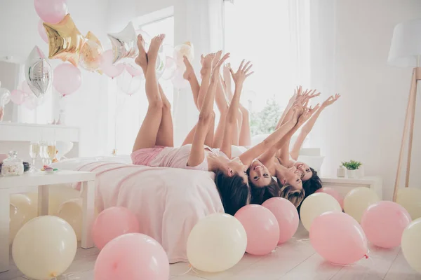 Profile side view of nice winsome lovely attractive cheerful cheery dreamy optimistic girlfriends having fun lying on bed raising legs hands up in light white interior decorated house