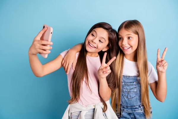 De cerca foto dos poco ella su rubia morena niñas largo bonito pelo teléfono hacer tomar selfies para mamá mamá espectáculo v-signo de usar casual jeans denim camisetas aislado azul brillante fondo —  Fotos de Stock