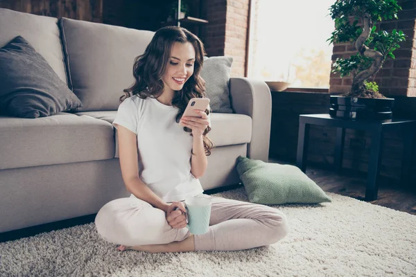 Close-up foto mooie brunette ze haar telefoon dame handen warme drank benen gekruiste armen schrijven brief dragen sportieve binnenlandse lichte kleur t-shirt broek kleding outfit sit vloer comfortabele divan — Stockfoto