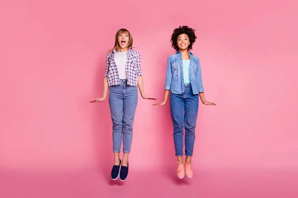 Full length body size view portrait two funky diversity she her ladies jumping high playful childish interested curious wear casual jeans denim checkered shirt clothes outfit isolated pink background