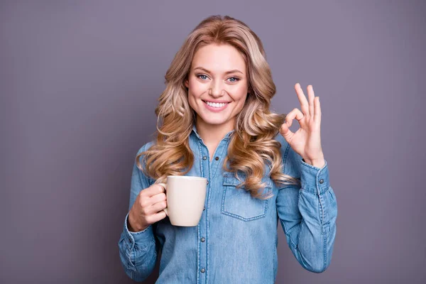 Retrato de agradável bonito encantador doce encantador alegre otimista alegre de cabelos ondulados senhora vestindo camisa azul segurando quente saboroso latte cacau cappuccino mostrando ok-sinal isolado sobre fundo cinza — Fotografia de Stock