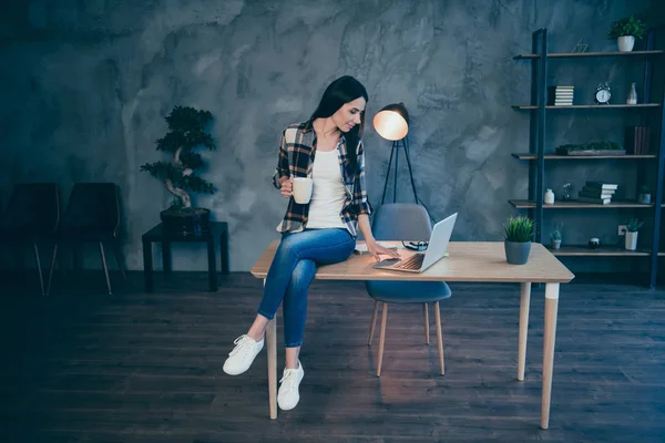 Full length body size view portrait of nice attractive charming cheerful brunette lady in checked shirt browsing news video chat sitting on desk at industrial loft style interior work place station — 图库照片