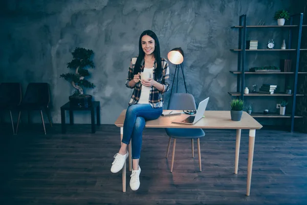 Longitud completa tamaño del cuerpo ver retrato de agradable atractivo encantador alegre alegre alegre morena dama en camisa a cuadros sentado en el escritorio tiempo libre en el estilo de loft industrial interior lugar de trabajo estación — Foto de Stock