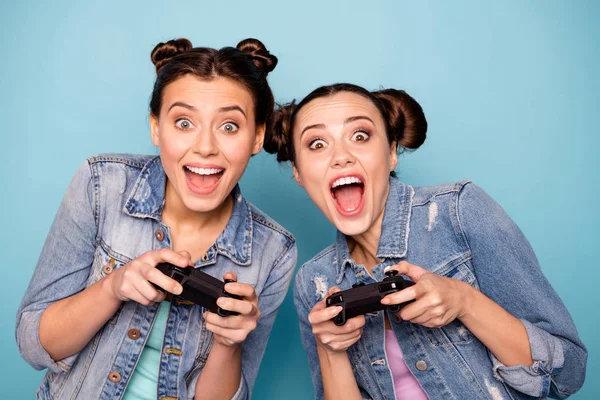 Закрыть фотографию симпатичных хипстеров, проводящих свободное время в игре Battle Speed Race с помощью геймпада Playstation, сидящих в помещении, одетых в джинсовый наряд на синем фоне — стоковое фото