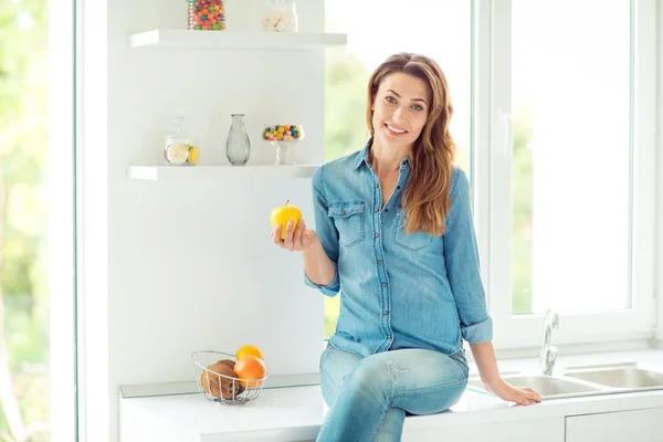 Portrait of her she nice-looking lovely charming cute attractive shine well-groomed cheerful brown-haired lady holding in hand apple sitting on countertop in light white interior style kitchen — Stock Photo, Image