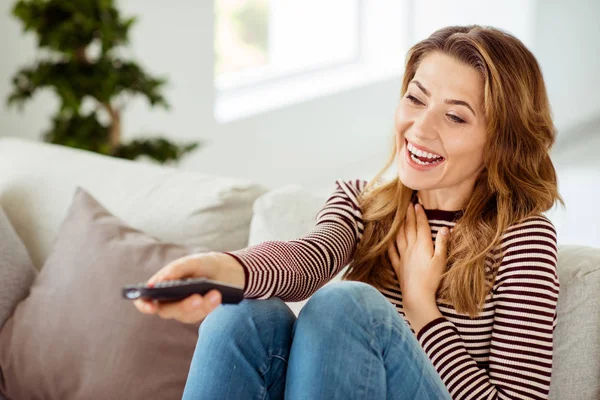 Close up side profile photo beautiful she her lady remote controller hands laughing loud change channels comic serial wear striped pullover clothes sit comfort divan house living room indoors — стоковое фото