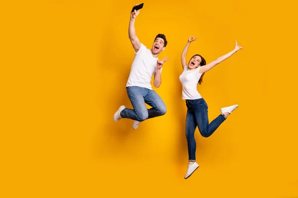 Full length side profile body size photo funky funny crazy people she her he he his guy lady jump high show v-sign make take selfies wear casual jeans denim white t-shirts isolated yellow background — стоковое фото