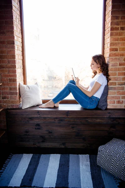 Vertikální pohled na její stranu je hezká atraktivní, krásně orientovlasá dívka, která sedí na okenní parapetu a čte si e-knihu z průmyslového podkroví ve stylu dřevěné cihly — Stock fotografie