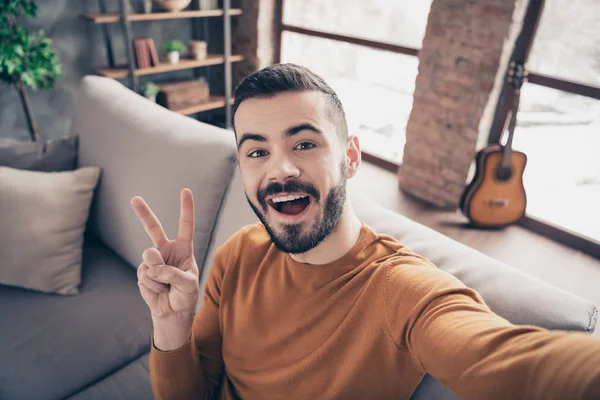 Закрыть фотографию красивого довольного парня-хипстера, сделать v-знаки смеха Свободное время одетый оранжевый пуловер свитер сидеть диван большой светлой комнате квартира vlog фото — стоковое фото