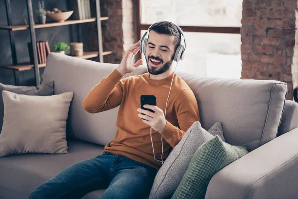 Retrato de hermoso hipster encantador estudiante uso dispositivo de tecnología moderna tiene música favorita cantar fuerte regocijo fin de semana jersey suéter jeans denim sentarse diván gran habitación moderna — Foto de Stock