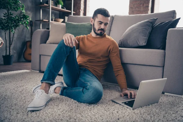 Retrato de agradable bastante concentrado encantador estudiante hipster desgaste zapatillas blancas vaqueros ver pantalla tecnología moderna conexión inalámbrica suéter de jersey naranja sentarse gran sala de luz interior — Foto de Stock