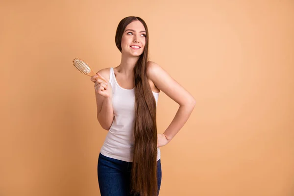 Close up foto hermosa su señora sostiene brazos manos cepillo de pelo mirada espacio vacío pelo marrón muy largo gran condición rizos usar casual blanco tank-top jeans denim aislado pastel beige fondo — Foto de Stock