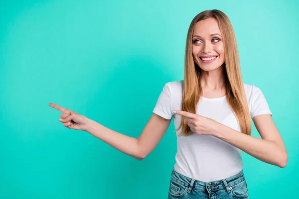 Portret van vrolijke promotor toont advertenties met haar wijsvinger dragen denim jeans geïsoleerd over Teal Turquoise achtergrond — Stockfoto