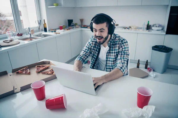 Portrait of his he nice attractive focused cheerful cheery guy wearing checked shirt attending online battle win winner in modern light white interior style kitchen