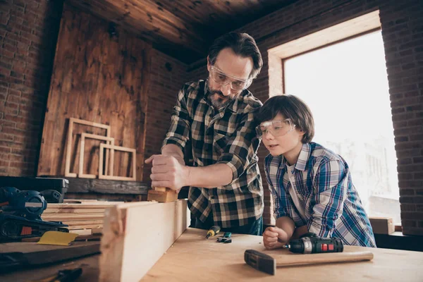 Portrait of focused serious pensive daddy son kid touch wood hold hand equipment craftsman fix engineer plank bearded checkered shirt brunet hair interior indoors glasses goggles protective