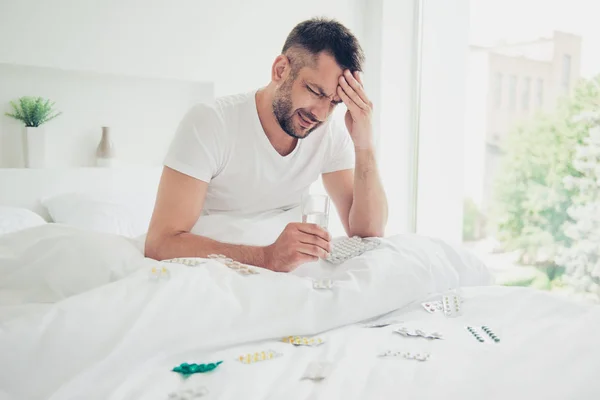 Close up photo amazing moody he him his unhappy guy hold hand arm head forehead glass water taking pills reduce strong sharp hurt pain early morning white sleep wear sit bedroom room house indoors