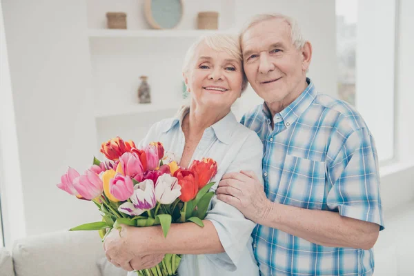 Foto de duas pessoas idosas adoráveis abraçando aniversário par surpresa grande vermelho tulipas bando apartamento dentro de casa — Fotografia de Stock