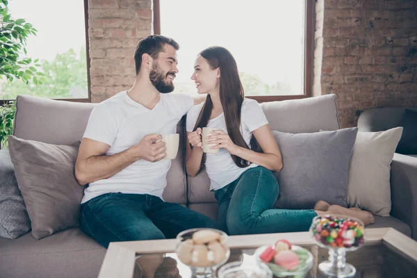 Foto de perfil de adorable pareja enamorada sosteniendo bebidas calientes manos mirada ojos sentado acogedor sofá interior — Foto de Stock