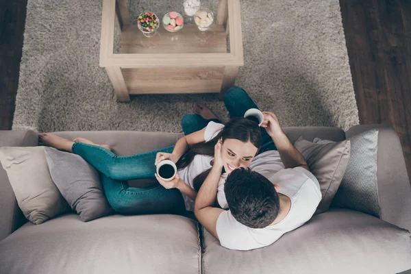 Foto de ángulo alto de adorable pareja enamorada sosteniendo las manos de bebidas calientes mira ojos alegres sentado sofá en el interior — Foto de Stock
