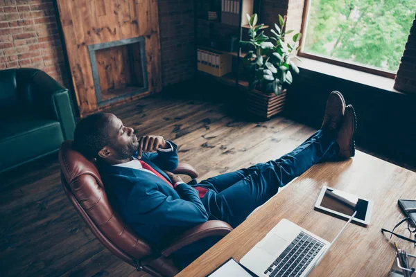 Dunkelhäutiger Typ, der verträumt durch das Fenster schaut und Beine auf dem Tisch hält, trägt elegantes Kostüm und sitzt im Bürostuhl — Stockfoto