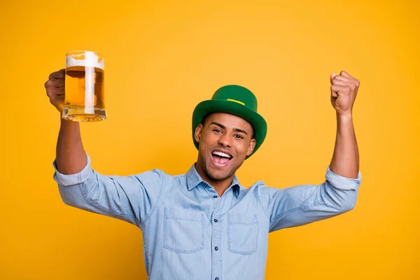 De cerca la foto del chico de piel oscura cogido de la mano levantar cerveza vidrio irlandés tradicional sombrero pub usar jeans camisa de mezclilla aislado fondo amarillo — Foto de Stock