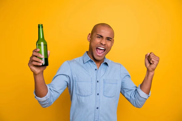 Foto de piel oscura macho levantar el puño botella de cerveza gritando fuerte meta sport bar usar jeans camisa de mezclilla aislado fondo amarillo brillante — Foto de Stock